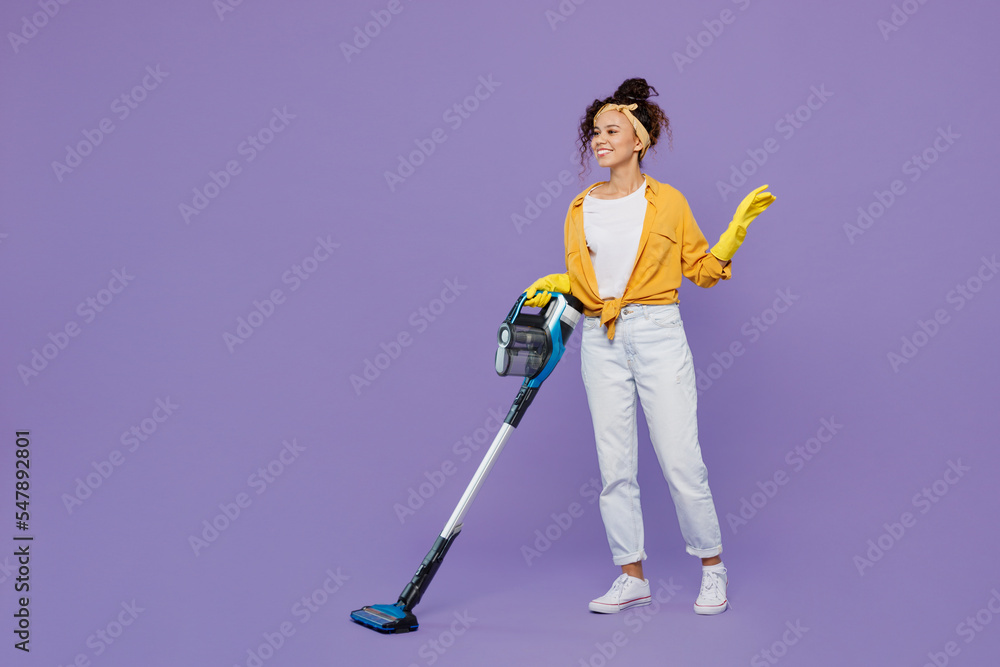 Full body side view smiling happy fun young housekeeper woman wearing yellow shirt rubber gloves use vacuum cleaner tidy up isolated on plain pastel light purple background studio. Housework concept. Full body side view smiling happy fun young housekeeper woman wearing yellow shirt rubber gloves use vacuum cleaner tidy up isolated on plain pastel light purple background studio. Housework concept.