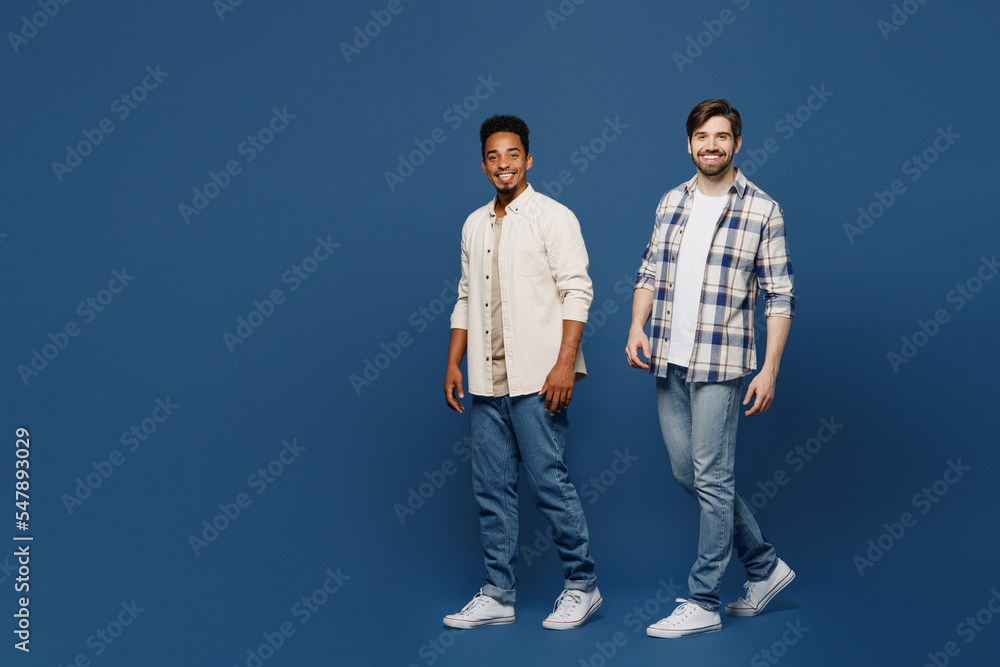 Full body side view young two friends happy men 20s wear white casual shirts together looking camera walking going strolling isolated plain dark royal navy blue background. People lifestyle concept.
