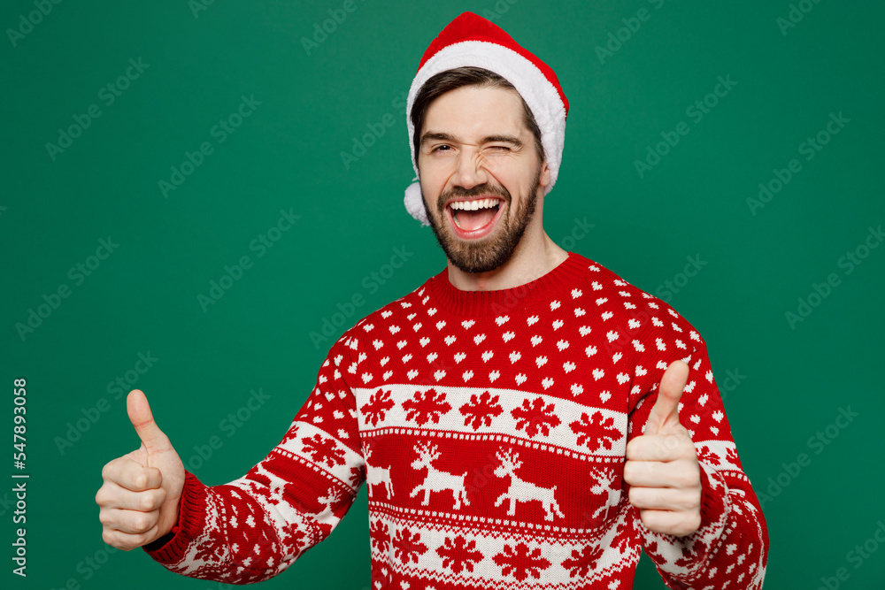 Merry fun young man wear red warm knitted sweater Santa hat posing showing thumb up like gesture wink isolated on plain dark green background studio portrait New Year 2023 holiday celebration concept Merry fun young man wear red warm knitted sweater Santa hat posing showing thumb up like gesture wink isolated on plain dark green background studio portrait New Year 2023 holiday celebration concept