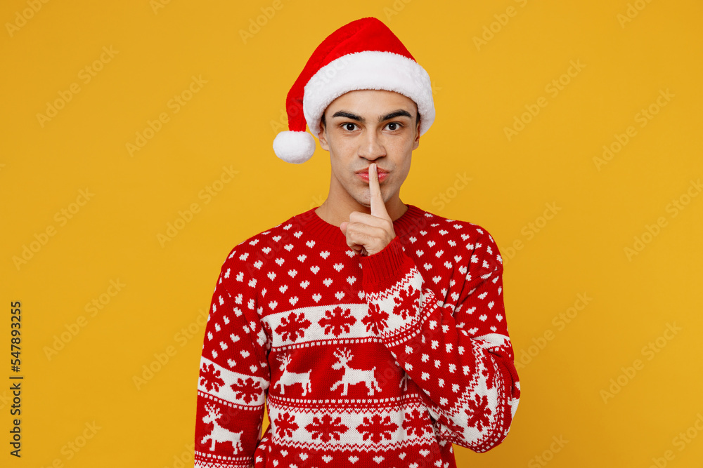 Merry young man wear knitted christmas sweater Santa hat posing say hush be quiet with finger on lips shhh gesture isolated on plain yellow background. Happy New Year 2023 celebration holiday concept. Merry young man wear knitted christmas sweater Santa hat posing say hush be quiet with finger on lips shhh gesture isolated on plain yellow background. Happy New Year 2023 celebration holiday concept.
