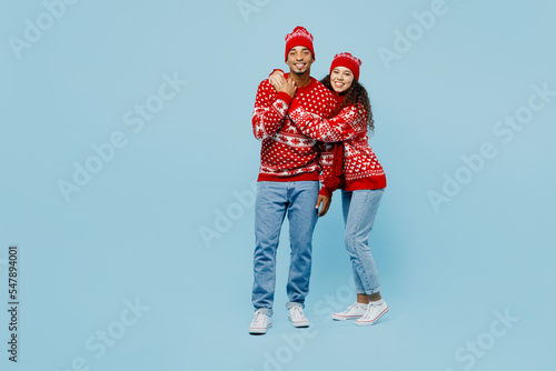 Full body merry young lovely smiling couple two man woman in red Christmas sweater Santa hat posing hug look camera isolated on plain pastel light blue background Happy New Year 2023 holiday concept