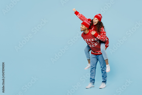 Full body joyful merry young couple two man woman in red Christmas sweater Santa hat posing give piggyback ride sit on back super hero gesture isolated on plain blue background New Year 2023 concept