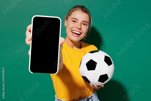 Young woman fan in basic yellow t-shirt cheer up support football sport team hold soccer ball watch tv live stream use mobile cell phone closeup blank screen isolated on dark green background studio © ViDi Studio