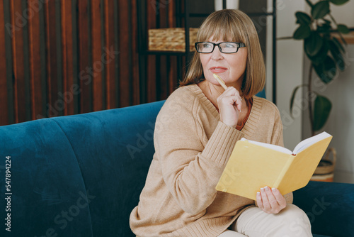 Elderly dreamful calm woman 50s year old in casual clothes sit on blue sofa write down in notebook diary look aside stay at home flat rest relax spend free spare time in living room indoor grey wall photo