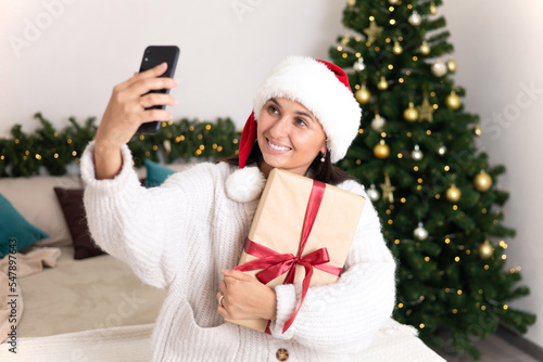Merry Christmas. A beautiful young woman in Santa's hat makes a video call to her family lying in the bedroom with a decorated Christmas tree and lights in the background. Lifestyle