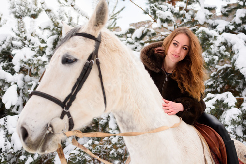 Winter walk on horses in the forest. A girl in a fur coat with a bright scarf in the snow. White and black colors. In the background are green Christmas trees and pine trees