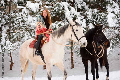 Winter walk on horses in the forest. A girl in a fur coat with a bright scarf in the snow. White and black colors. In the background are green Christmas trees and pine trees © Iryna Savchuk