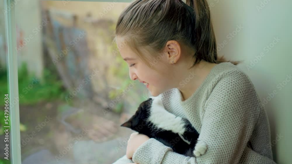 Cute handsome domestic black cat licks cheeks of happy smiling girl sitting at home window on windowsill during sunset. Happy carefree childhood concept Cute handsome domestic black cat licks cheeks of happy smiling girl sitting at home window on windowsill during sunset. Happy carefree childhood concept