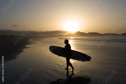 Silueta de hombre caminando por la playa con una tabla de surf en un atardecer de verano en la playa de Famara en Lanzarote photo