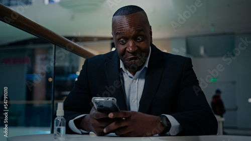 African American adult middle-aged mature man employee businessman indoors in office cafe table looking phone typing chatting texting mobile message online business chat browsing smartphone thinking