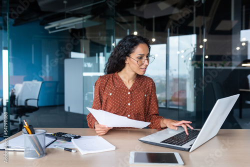 Canvas Print Young happy and successful businesswoman in glasses working with documents inside office, Hispanic woman with laptop looking at bills and contracts, financier with curly hair using laptop
