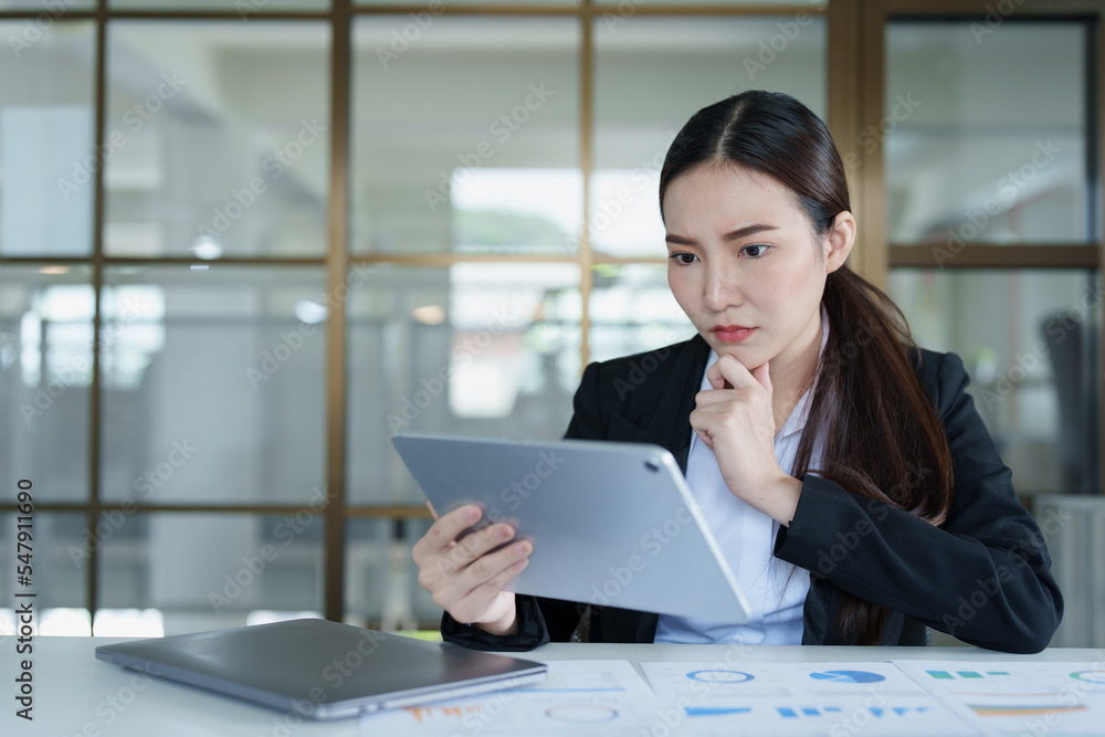 Portrait of a thoughtful Asian businesswoman looking at financial statements and making marketing plans using a tablet computer on her desk Portrait of a thoughtful Asian businesswoman looking at financial statements and making marketing plans using a tablet computer on her desk