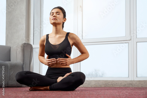 Front view of beautiful girl practicing yoga pose with closed eyes indoors. Brunette slim female sitting barefoot on crossed legs holding by hands. Concept of yoga practicng