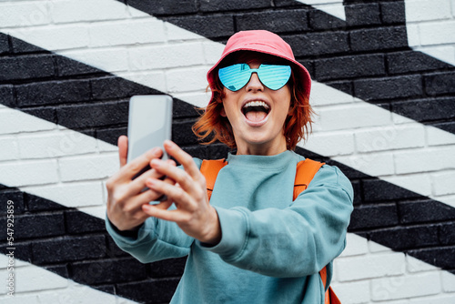 Excited redhead woman screaming while taking a selfie photo outdoors. Emotional hipster fashion women in bright clothes, heart shaped glasses, bucket hat taking selfie photo on the phone camera photo