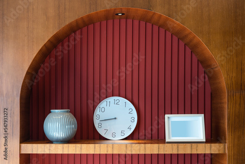 Obraz na plátně Modern clock, white photo frame, blue ceramic vase on the wooden shelf built-in of the red wall stripes decoration for interior home and living of office building