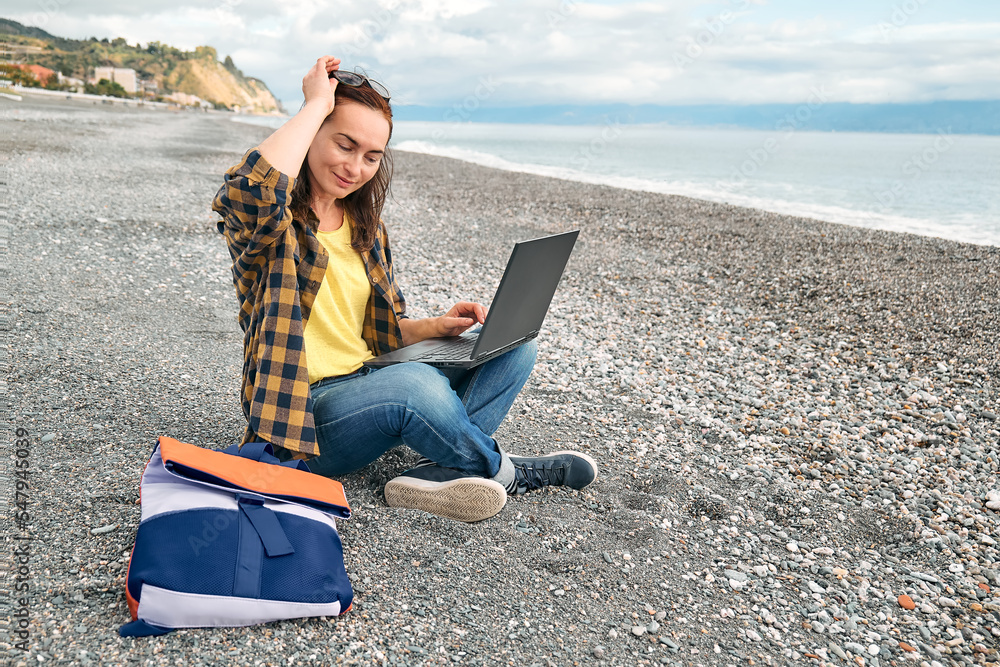 Woman in warm plaid shirt with laptop computer working outdoors while sitting on winter beach in front of the sea. Modern lifestyle, connection, blogging, business, freelance work.