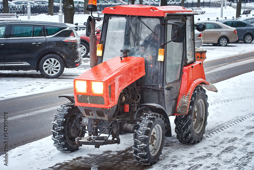 Tractor with mounted salt spreader ready to work on snowy sidewalk, snow removal service operating in winter during blizzard. Snow and ice removal service spread rock salt on road and walkway.