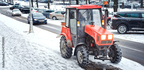 Red tractor with salt spreader ready to work on snowy sidewalk, snow removal service operating in winter during blizzard. Snow and ice removal service spread rock salt on road and walkway, melt snow