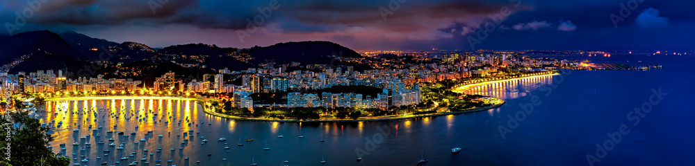 Panoramic photograph of the shore of Guanabara Bay in Rio de Janeiro at night with the buildings and citylights in Botafogo and Flamengo neighborhoods and the hills in the background