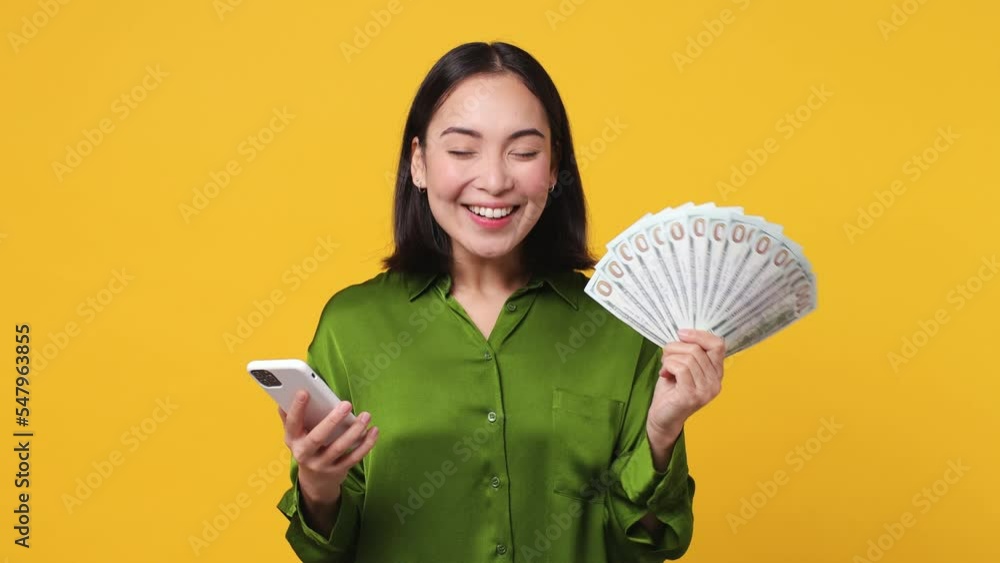 Excited happy joyful young woman of Asian ethnicity 20s she wear green shirt use mobile cell phone hold win fan of cash money in dollar banknotes isolated on plain yellow color wall background studio Excited happy joyful young woman of Asian ethnicity 20s she wear green shirt use mobile cell phone hold win fan of cash money in dollar banknotes isolated on plain yellow color wall background studio