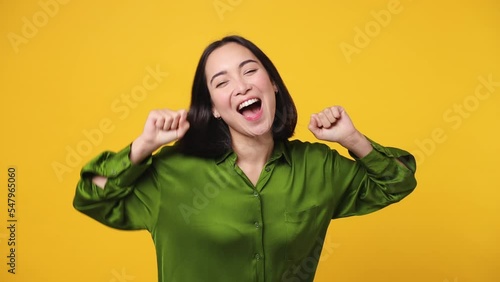 Beautiful fancy vivid happy bright young woman of Asian ethnicity she wear green shirt dance rising have fun enjoy relax celebrate play fluttering hair isolated on plain yellow color wall background