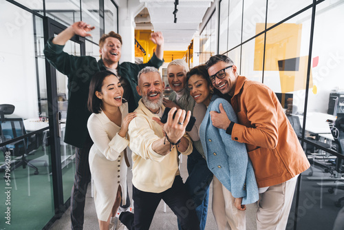 Fotografia Group of cheerful multiethnic friends taking selfie on smartphone near glass wal
