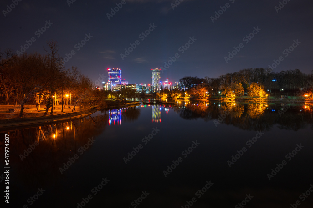 Bukarest - Parklandschaft mit See bei Nacht mit modernen beleuchteten Bürogebäuden im Hintergrund