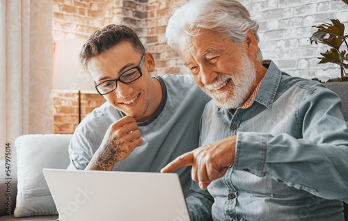 Senior man and young boy with tattoo smiling and looking together at laptop at home. Happy nephew teaching and showing new computer technology to his old grandfather. OurMindsMatter photo