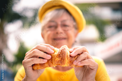 Blurred elderly woman having a relaxing moment at the outdoor cafe breaking her croissant in half. Attractive Caucasian older lady in yellow enjoying a fragrant croissant