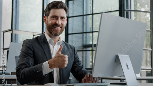 Middle-aged adult bearded 40s businessman worker working with computer in office showing thumb up. Manager man boss male company CEO typing working online show recommend sign approval gesture agree
