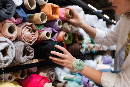 cropped view of salesperson choosing between colorful fabric rolls on shelves of textile shop