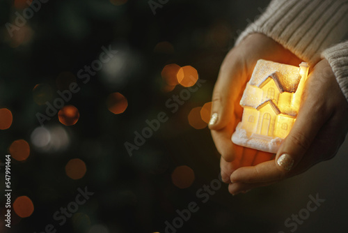Hands holding illuminated little house on background of christmas tree with lights bokeh. Cozy home. Atmospheric Holiday banner. Family and safety concept. Merry Christmas!