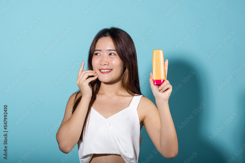 Beautiful Asian young woman holding body lotion on hand in studio shot isolated on blue background, Smiling brunette female hold cream sunblock bottle, tube of sunscreen, protect sun light and UV Beautiful Asian young woman holding body lotion on hand in studio shot isolated on blue background, Smiling brunette female hold cream sunblock bottle, tube of sunscreen, protect sun light and UV