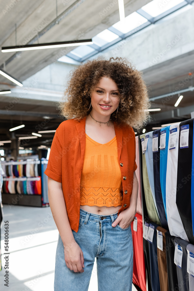 curly woman standing with hand in pocket and smiling at camera in textile shop - obrazy, fototapety, plakaty 