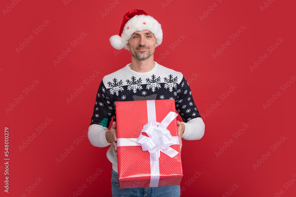 Optimistic man in Santa hat and winter sweater smiling and holding presents gift while celebrating Christmas against red background. Optimistic man in Santa hat and winter sweater smiling and holding presents gift while celebrating Christmas against red background.