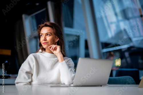 Thinking middle aged business woman using laptop while sitting at desk at the office. Executive business woman wearing sweater. 