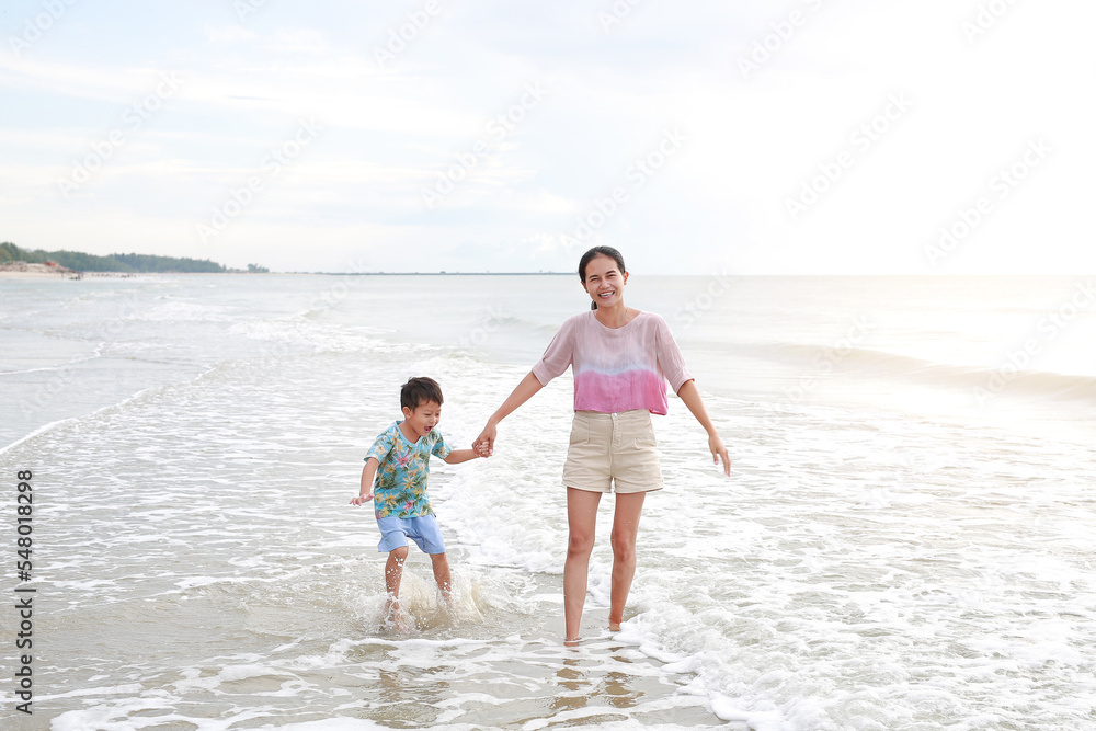 Happy Asian mom and son enjoy playing on tropical sand beach at sunrise. Happy family in summer holiday.