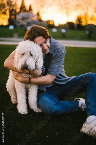 Teenage boy hugs golden doodle dog in front of sunset trees photo