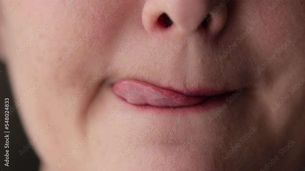 Young woman licks pink lips with tongue and bites lower lip with teeth. Part of female face with chin and mouth on dark background extreme closeup Young woman licks pink lips with tongue and bites lower lip with teeth. Part of female face with chin and mouth on dark background extreme closeup