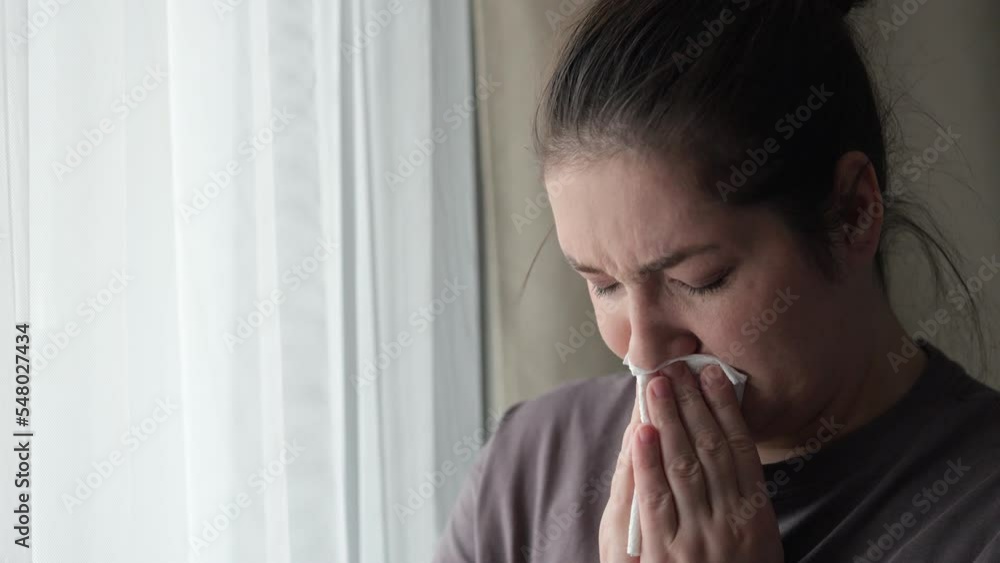 Sick brunette woman sneezes covering nose with napkin feeling unwell. Housewife tries to handle cold standing near bright window in bedroom closeup Sick brunette woman sneezes covering nose with napkin feeling unwell. Housewife tries to handle cold standing near bright window in bedroom closeup