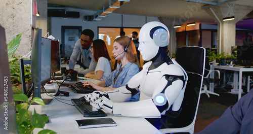 Robot working at computer among people. Maschine typing on keyboard in office. IT team of future. Futuristic worker. Humanoid work at call center. Support job. Selling concept. Technologies. photo