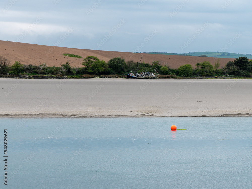 View of Clonakilty Bay at low tide. A small orange buoy is kept on a calm water surface. The sandy shore of the sea bay. The coastline on a cloudy day. Seaside landscape.