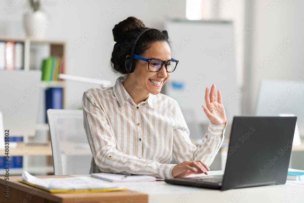 Excited female office worker making video call via laptop, talking to webcam and waving, sitting at workplace in office Excited female office worker making video call via laptop, talking to webcam and waving, sitting at workplace in office