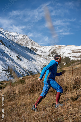 Dressed up man in sportswear hiking a grassy slope. In warm wind resistant clothes. Next to a snowy mountain.