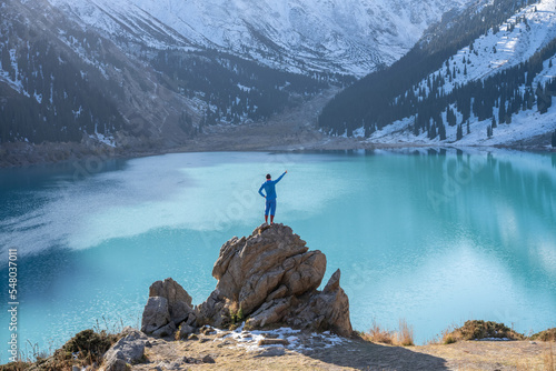 Looking at a view man standing on a huge boulder on a vantage point over stunning turquoise lake surrounded by snowy mountains on a sunny day. His hands are outstretched like wings, facing camera.