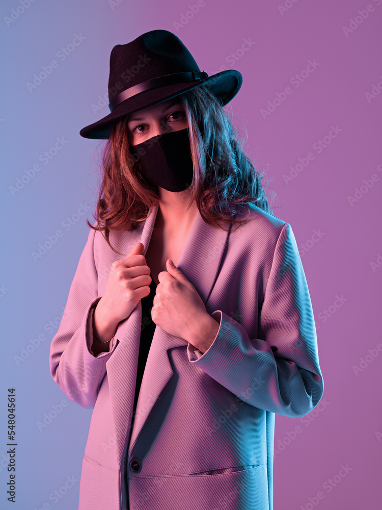 Surprised fashionable young woman in a coat, hat and protective mask in purple neon lighting.