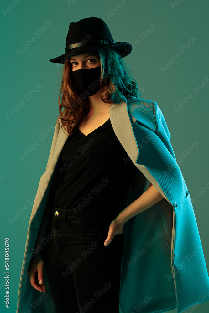 Portrait of a trendy young woman in a coat, hat and protective mask on a green background.