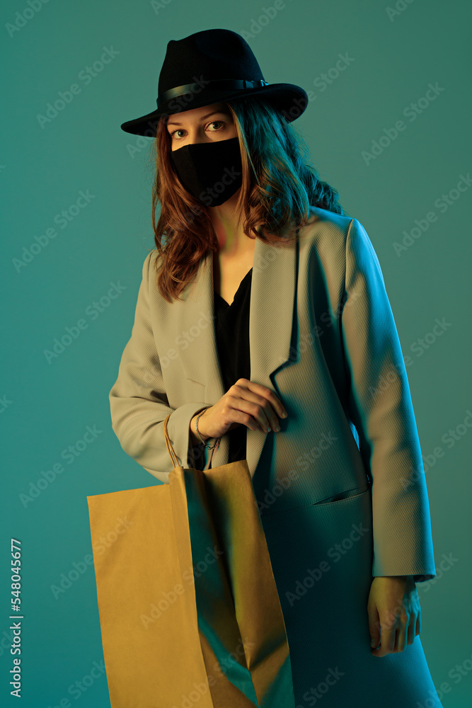 Portrait of a trendy young woman in a coat, hat and protective mask with a paper shopping bag on a green background.