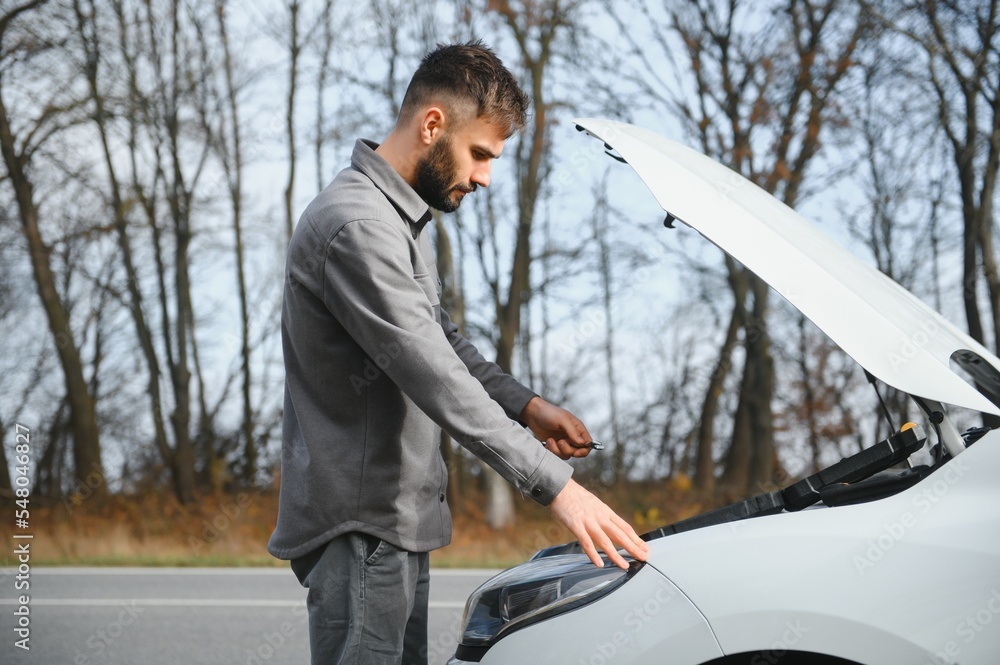 A young man near a broken car with an open hood on the roadside.