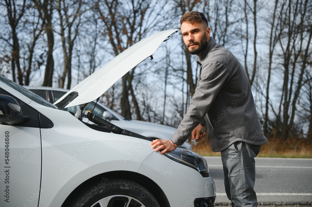 Young upset casual man trying to fix his broken car outdoors. Man waiting for towing service for help car accident on the road. Roadside assistance concept.